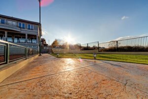 A baseball field under a bright sun, with clouds above and stamped concrete surrounding the area.